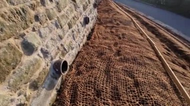 Çimento karıştırıcısının uzun düz duvarı. Duvarın arkasında duvarın içinden drenaj borularıyla çıkarılan toprak var. Kanalizasyon borusu çekiliyor. İnşaat alanı, mezarlık, gabion.