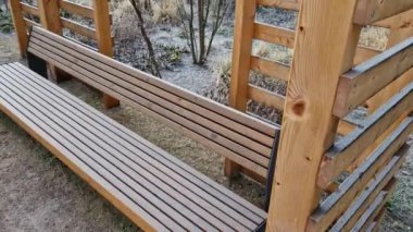 Duvarları ve çatısı çizgili ahşap pergola. Tırmanma bitkileri için çardak görevi görüyor. Kumlu bej yüzey. Tel banklarla parktaki patikalar. Vidaları ve iplikleri olan temel çubuklar, kış