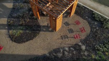 Duvarları ve çatısı çizgili ahşap pergola. Tırmanma bitkileri için çardak görevi görüyor. Kumlu bej yüzey. Tel banklarla parktaki patikalar. Vidaları ve iplikleri olan temel çubuklar, kış