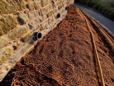 Çimento karıştırıcısının uzun düz duvarı. Duvarın arkasında duvarın içinden drenaj borularıyla çıkarılan toprak var. Kanalizasyon borusu çekiliyor. İnşaat alanı, mezarlık, gabion.