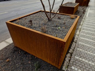 Çeşitli yükseklik ve şekillerdeki metal kaplar kaldırımı tuğla bina cephesinden işlek yoldan ayırıyor. Çalılar, çakıllar, mozaikler ve katmanlı saksılarda çalılarla daimi ve süslü otlar.