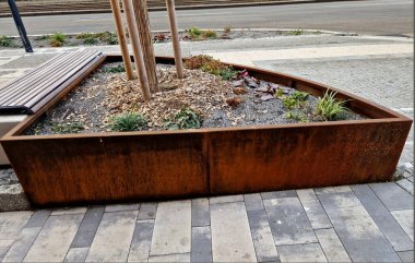 Çeşitli yükseklik ve şekillerdeki metal kaplar kaldırımı tuğla bina cephesinden işlek yoldan ayırıyor. Çalılar, çakıllar, mozaikler ve katmanlı saksılarda çalılarla daimi ve süslü otlar.