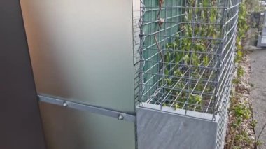 Gabion teli sepeti, kafesler. Doğal endüstriyel görünümüyle ön bahçede çit var. Galvanize kablodan yapılmış. Katlanmış taşlarla dolu istinat duvarı. Gürültü bariyeri yerleşim alanını koruyor.