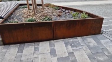 Çeşitli yükseklik ve şekillerdeki metal kaplar kaldırımı tuğla bina cephesinden işlek yoldan ayırıyor. Çalılar, çakıllar, mozaikler ve katmanlı saksılarda çalılarla daimi ve süslü otlar.