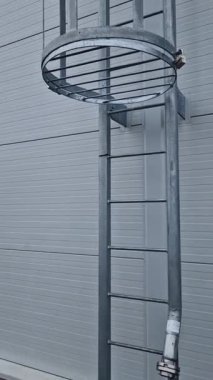 Koridorun çatısına çıkan yangın merdiveni koruyucu ızgaranın alt kısmındaki ızgarayla korunuyor. Beyaz metal depo duvarları. Galvanize demir ve yangın hortumunu bağlamak için bir boru.