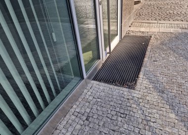 Binanın girişindeki endüstriyel paspas temizleme bölgeleri. Kemer ya da yarım daire şeklinde siyah plastik bir minder kireçtaşı mozaik fayansların üzerinde yatar.