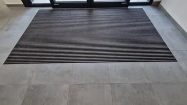 Binanın girişindeki endüstriyel paspas temizleme bölgeleri. Kemer ya da yarım daire şeklinde siyah plastik bir minder kireçtaşı mozaik fayansların üzerinde yatar.