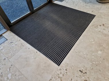 Binanın girişindeki endüstriyel paspas temizleme bölgeleri. Kemer ya da yarım daire şeklinde siyah plastik bir minder kireçtaşı mozaik fayansların üzerinde yatar.