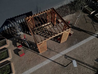 wooden pergola with striped planks on walls and roof. serves as a trellis for climbing plant. sandy beige surface. paths in park with wire benches. foundation rods with screws and threads, sunlight clipart