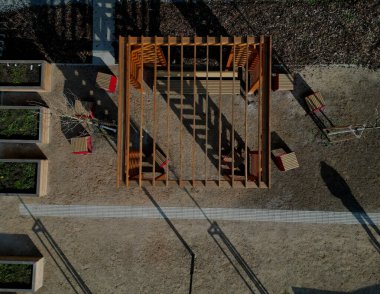 Duvarları ve çatısı çizgili ahşap pergola. Tırmanma tesisi olarak kullanılıyor. Kumlu bej yüzey. Tel banklarla parktaki patikalar. Temelde vida, iplik ve güneş ışığı olan çubuklar