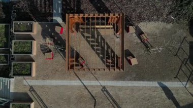 Duvarları ve çatısı çizgili ahşap pergola. Tırmanma tesisi olarak kullanılıyor. Kumlu bej yüzey. Tel banklarla parktaki patikalar. Temelde vida, iplik ve güneş ışığı olan çubuklar