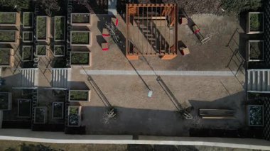 Duvarları ve çatısı çizgili ahşap pergola. Tırmanma tesisi olarak kullanılıyor. Kumlu bej yüzey. Tel banklarla parktaki patikalar. Temelde vida, iplik ve güneş ışığı olan çubuklar