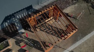 Duvarları ve çatısı çizgili ahşap pergola. Tırmanma tesisi olarak kullanılıyor. Kumlu bej yüzey. Tel banklarla parktaki patikalar. Temelde vida, iplik ve güneş ışığı olan çubuklar