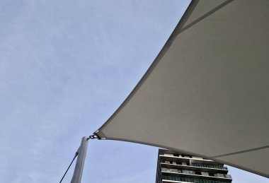 Beyaz tente gergin, güneşten gölgeleniyor ve restoranın terasında yağmur yağıyor, çocuk parkında, platformda. Direk ve gergin halatlar uçlarına sıkıca bağlanmış.