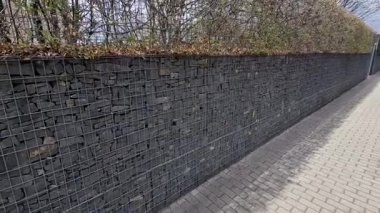 Bir apartman bloğundaki bahçeler arasında kıvrımlı duvarlar. Çakıl parkı yüzeyi olan halka açık bir yola açılan metal kapılar. Gabion sepetlerinde istiflenmiş taşlar
