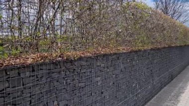 Bir apartman bloğundaki bahçeler arasında kıvrımlı duvarlar. Çakıl parkı yüzeyi olan halka açık bir yola açılan metal kapılar. Gabion sepetlerinde istiflenmiş taşlar