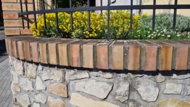 Metal parmaklıklı çitli evin yanındaki duvara bakıyorum. Bahçe bahçesi alçak çiçeklerle kaplı. Klasik tuğla ve taşların birleşimi.