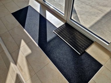 Binanın girişindeki endüstriyel paspas temizleme bölgeleri. Kemer ya da yarım daire şeklinde siyah plastik bir minder kireçtaşı mozaik fayansların üzerinde yatar.