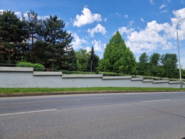 Yolun kenarındaki borular parmaklıklı büyük beton istinat duvarı. Arka planda çiçekler açan çalılar, ağaçlar ve park