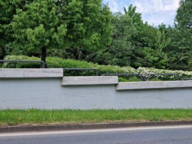 Yolun kenarındaki borular parmaklıklı büyük beton istinat duvarı. Arka planda çiçekler açan çalılar, ağaçlar ve park