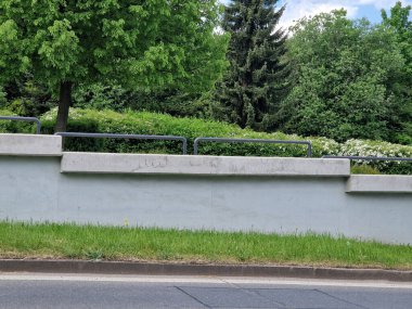 Yolun kenarındaki borular parmaklıklı büyük beton istinat duvarı. Arka planda çiçekler açan çalılar, ağaçlar ve park