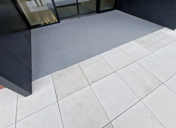 Keçeli mendil, kaset fırçası, gri alüminyum. Profiller asfalt seviyesinin altına gömülmüş. Değiştirilebilir keçeli kalemler ayakkabıları temizler ve suyu emer. Kayma riski azaldı.