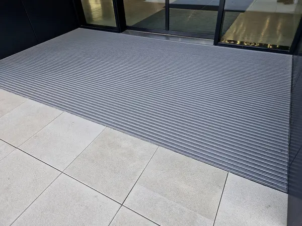 Keçeli mendil, kaset fırçası, gri alüminyum. Profiller asfalt seviyesinin altına gömülmüş. Değiştirilebilir keçeli kalemler ayakkabıları temizler ve suyu emer. Kayma riski azaldı.