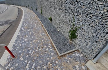 Yolun yamacını tutan çirkin beton duvar doğal taşlarla dolu bir gabionla kaplı. Astarın görünümü daha doğal.