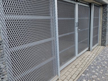 Ev atıklarını ayırmak için konteynır yerleştirme alanı. Gri kapı ve delinmiş metal ızgaranın yarısı, genişletilmiş metal. Taş kablolar dolu, otopark ofisi dolu. 