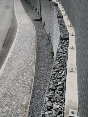 Yolun yamacını tutan çirkin beton duvar doğal taşlarla dolu bir gabionla kaplı. Astarın görünümü daha doğal.