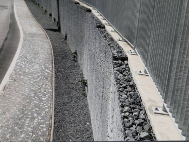 Yolun yamacını tutan çirkin beton duvar doğal taşlarla dolu bir gabionla kaplı. Astarın görünümü daha doğal.