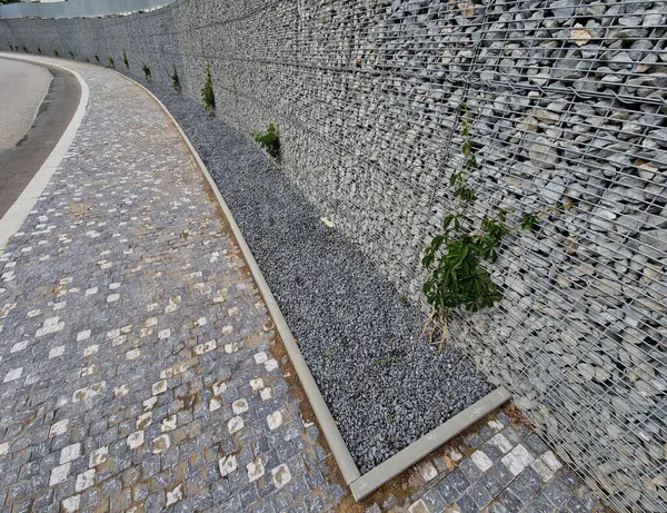 Yolun yamacını tutan çirkin beton duvar doğal taşlarla dolu bir gabionla kaplı. Astarın görünümü daha doğal.