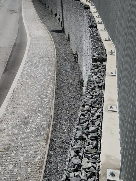 Yolun yamacını tutan çirkin beton duvar doğal taşlarla dolu bir gabionla kaplı. Astarın görünümü daha doğal.