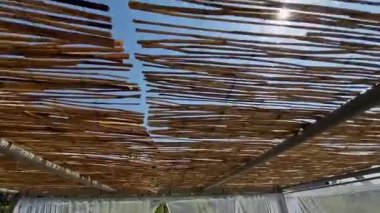 İpek kumaş, metal borulardan yapılmış bir pergola üzerinde çırpınır. Bu perde bir duvağa ya da gelinliğe benziyor. Rüzgâr, şeffaf kumaşları deniz kıyısında terasın üzerinde hareket ettiriyor.