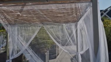 İpek kumaş, metal borulardan yapılmış bir pergola üzerinde çırpınır. Bu perde bir duvağa ya da gelinliğe benziyor. Rüzgâr, şeffaf kumaşları deniz kıyısında terasın üzerinde hareket ettiriyor.