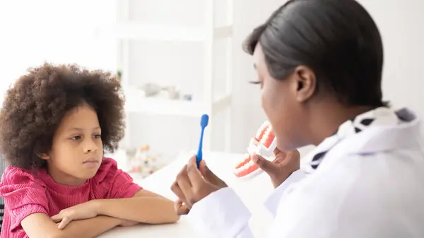 African American dentist explaining to mixed-race afro child how to brush teeth. The doctor and healthcare concept for children.
