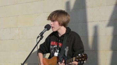 Bender, Moldova - November 12, 2022: Teen-ager Guy playing guitar on street and sing. A young guy of European appearance with blond hair plays the guitar on the street and sings on a sunny day