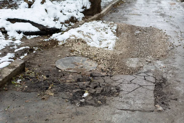 Tamamlanmamış yol onarımı. Kanalizasyon kapağı ve kırılmış asfalt parçalanmış taşlarla kaplı. Yerleşim alanındaki bozuk bir yolda toprak ve kar. Sığ alan derinliği, seçici odak