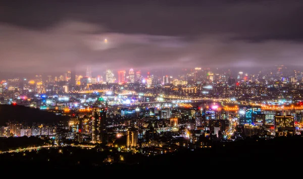 Clouds moving in the sky at night. Neon lights shine on the vibrant cityscape. Night view of the city surrounded by mountains is hazy and dreamy. Taipei City, Taiwan