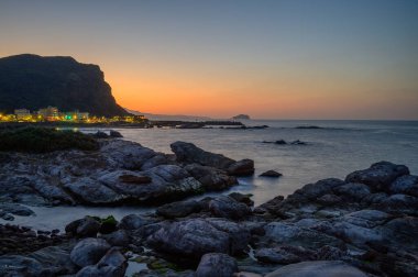 Deniz kıyısındaki bir köy alacakaranlıkta turuncu bir gökyüzüne karşı parlıyor. Ruifang Nanya, New Taipei City 'nin tuhaf kayalık manzarası. Tayvan