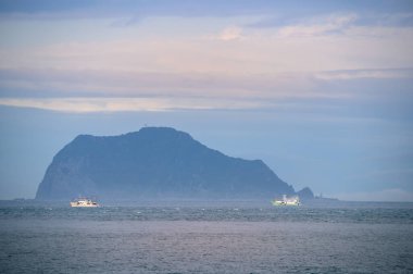 Tekne adanın etrafında döner. Biraz sis ve renkli bulutlar var. New Taipei 'deki Wanli Sahili' nden Keelung Adası manzarası. Tayvan.