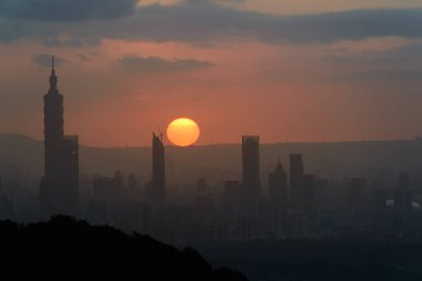 Gün batımında, güneş bulutların arasından doğar ve Taipei şehrinde parlar. Şehir binalarının siluetleri. Turuncu gökyüzü ve kara bulutlar.