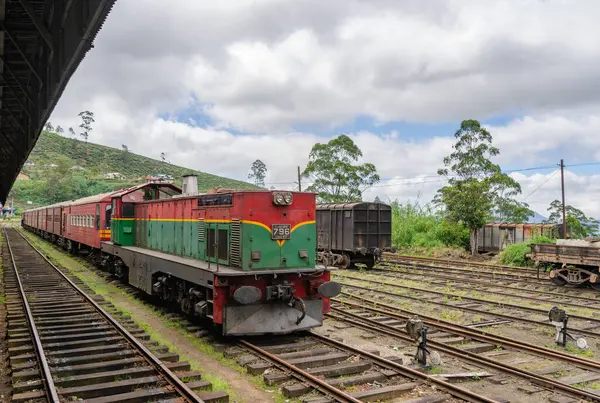 A vibrant green and red train rests on the tracks, ready for departure. The lush green hills and cloudy sky paint a serene backdrop. Captured on 2024-01-28, Ambewela, Sri Lanka.
