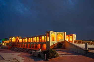 Tarihi Khaju Köprüsü, alacakaranlıkta derin mavi gökyüzünün altında parlak turuncu ışıklarla parlar. Pers mimarisinin sembolü, İsfahan, İran.