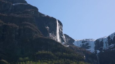 Yüksek bir şelale engebeli bir uçurumdan aşağı yuvarlanır ve arka planda açık mavi bir gökyüzü vardır. Kar, Stegstein, Norveç yakınlarındaki kontrastı artırıyor..