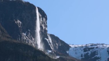 Yüksek bir şelale engebeli bir uçurumdan aşağı yuvarlanır ve arka planda açık mavi bir gökyüzü vardır. Kar, Stegstein, Norveç yakınlarındaki kontrastı artırıyor..