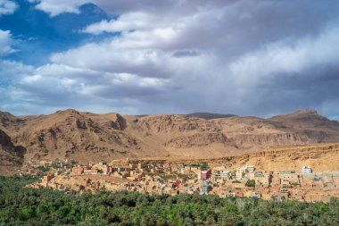 Lush palm trees surround mud-brick buildings against towering cliffs under a dynamic sky. Captured in Todra Gorges, Morocco. clipart