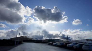 Mavi gökyüzünün altında dağınık bulutlarla jeotermal alandan beyaz buhar yükseliyor. Park halindeki arabalarda insan varlığı öneriliyor. Reykjanes Yarımadası, İzlanda.