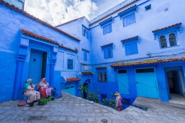 Chefchaouen, Fas - 25 Mart 2024: Mavi boyanmış binaların arasında bir kapı ağzında dinlenen yerli kadınlar. Kasabanın eşsiz mavi ve beyaz mimarisi büyüleyici ve rüya gibi bir atmosfer yaratır..