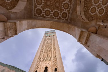 Hasan II Camii 'nin yükselen minaresinin alçak açılı görüntüsü, süslü desenli kemerleriyle bulutlu bir gökyüzüne karşı duruyor. Kazablanka, Fas.